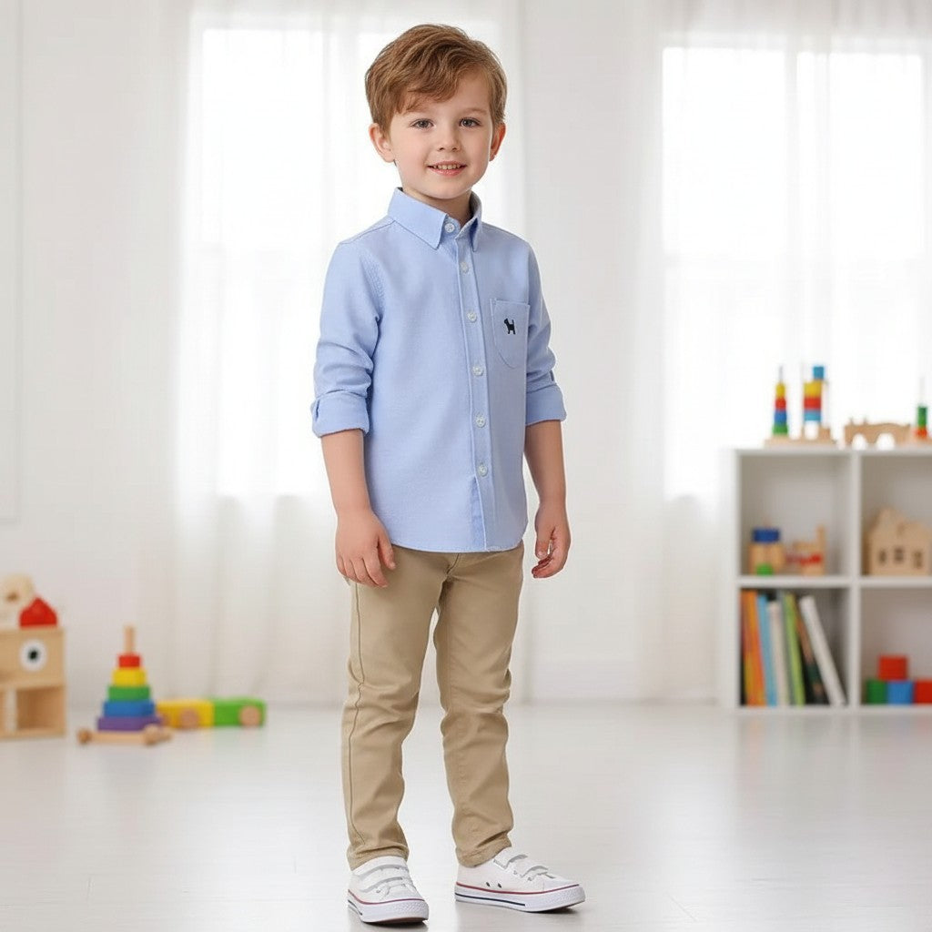 A young boy wearing a GIGI & POPO light blue long-sleeved button-down shirt with khaki trousers and white sneakers.